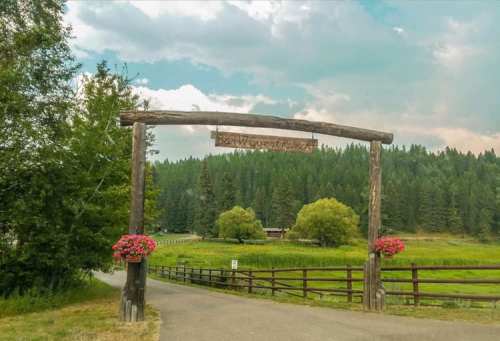 Wooden archway sign at a scenic entrance, surrounded by trees and flowers, leading to a lush green landscape.