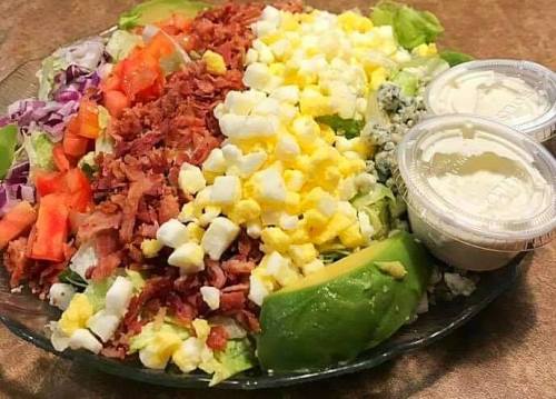 A colorful salad with layers of lettuce, tomatoes, bacon, cheese, avocado, and two containers of dressing.