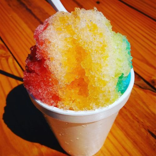 A colorful snow cone in a white cup, featuring layers of red, orange, yellow, green, and blue shaved ice.