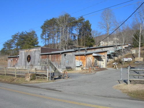 A rustic building with wooden siding, surrounded by trees, along a gravel road on a clear day.