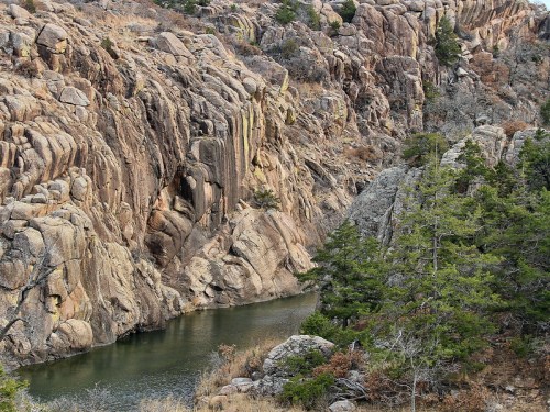 Rocky cliffs flank a calm river, with scattered trees and rugged terrain in a natural landscape.