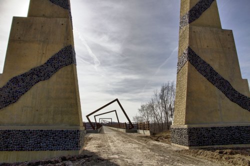 A pathway leads through two tall, textured pillars towards a geometric structure in the distance under a cloudy sky.