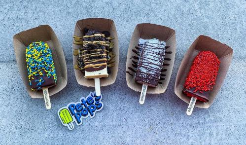 Four colorful ice cream pops on paper trays, topped with sprinkles, chocolate drizzle, and various toppings.