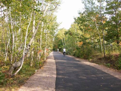 Two cyclists ride along a paved path surrounded by trees in a scenic, wooded area.