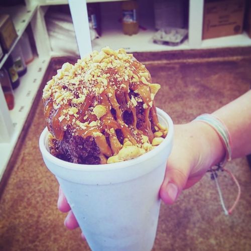 A hand holds a white cup filled with a large dessert topped with caramel and crushed peanuts.
