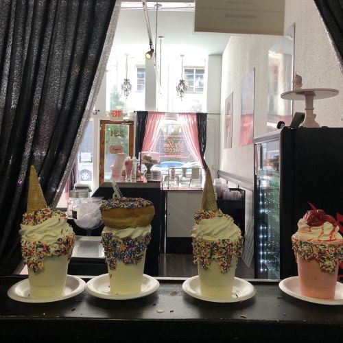 Four colorful ice cream sundaes with sprinkles and cones, displayed in a shop with a cozy interior in the background.
