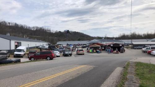 A bustling outdoor market with vendors, cars parked along the road, and hills in the background under a cloudy sky.
