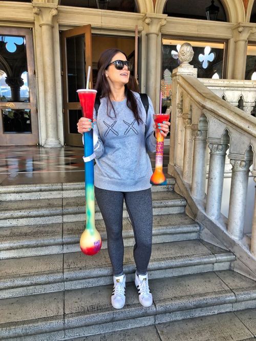 A woman in a gray sweater and leggings holds two colorful drinks while standing on a staircase.
