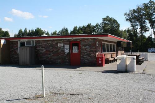 A small brick building with a red roof, surrounded by gravel, featuring a sign and outdoor seating. Trees in the background.