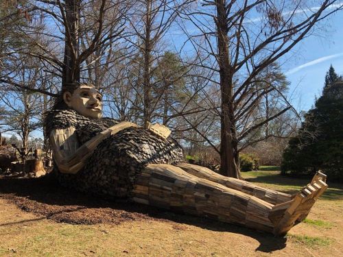 A large wooden sculpture of a reclining figure surrounded by trees in a park setting.