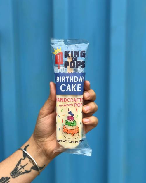 A hand holds a King of Pops Birthday Cake popsicle against a blue background. The packaging features colorful graphics and text.