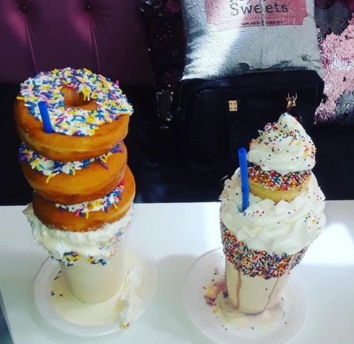 A tall milkshake topped with whipped cream and a donut, alongside a stack of three colorful donuts with sprinkles.