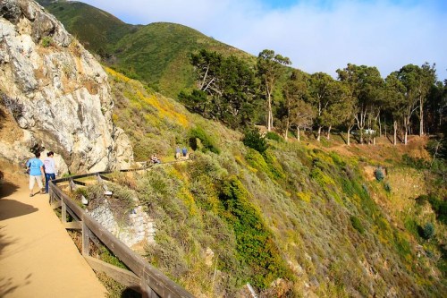 A scenic trail along a hillside with lush greenery and people walking, surrounded by mountains and blue sky.