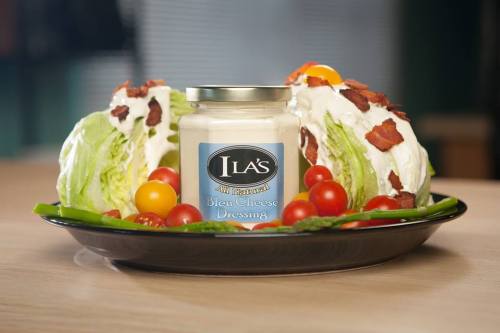 A jar of bleu cheese dressing surrounded by lettuce wedges, cherry tomatoes, and bacon on a black plate.