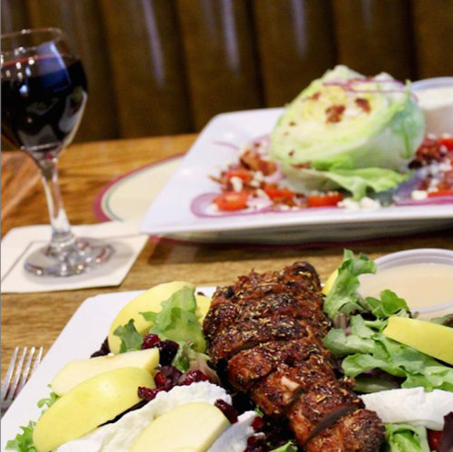 A plate of grilled chicken on a bed of greens with apples and cranberries, alongside a glass of red wine.