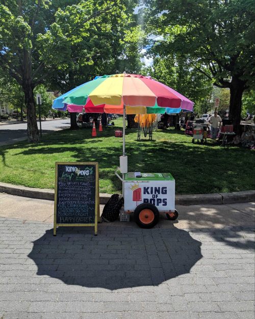 A colorful umbrella shades a popsicle cart with a chalkboard menu in a park surrounded by trees.