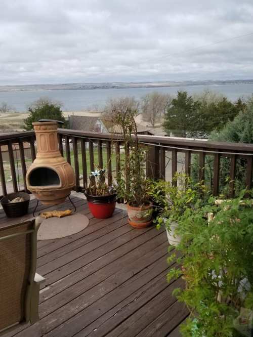 A wooden deck with potted plants and a clay chiminea, overlooking a lake and cloudy sky in the background.