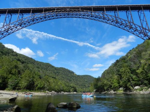 A scenic view of a river with a large bridge overhead, surrounded by lush green hills and a clear blue sky.