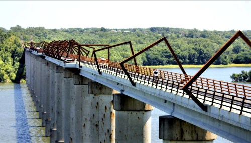 A unique bridge with angular metal supports spans a river, surrounded by lush greenery under a clear blue sky.