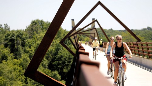 Two cyclists ride on a modern bridge surrounded by greenery, with geometric metal structures framing the scene.