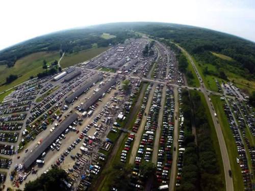Aerial view of a large parking lot filled with numerous cars, surrounded by green fields and trees.