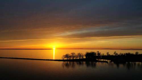 A serene sunset over a calm lake, with silhouettes of trees along the shore and vibrant orange and yellow hues in the sky.