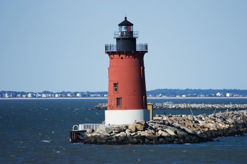 A red and white lighthouse stands on a rocky jetty by the water, with a clear blue sky in the background.