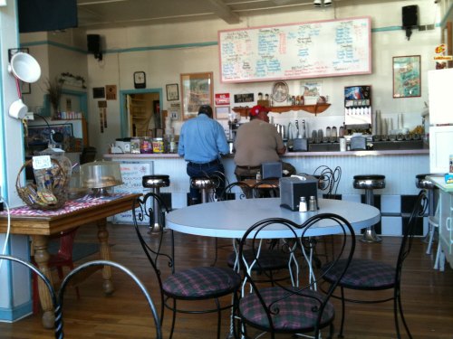 A cozy diner interior with two patrons at the counter, tables set for dining, and a menu board on the wall.