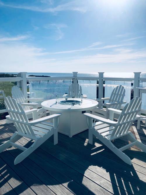A sunny outdoor seating area with white Adirondack chairs around a circular table, overlooking a scenic ocean view.