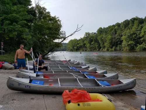 Two people by a row of canoes on a riverbank, surrounded by trees and calm water.