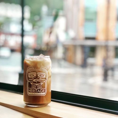 A glass of iced coffee with cream sits on a wooden table by a window, with a blurred outdoor view in the background.