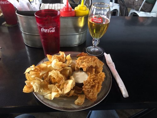 A plate of crispy fried chicken with potato chips and dipping sauce, accompanied by a glass of beer and a soda.
