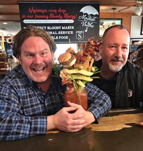 Two men smiling at a bar, one holding a towering Bloody Mary topped with food items, showcasing a fun dining experience.