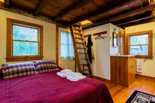 Cozy bedroom with a red quilt, wooden beams, a ladder to a loft, and a small bathroom area with towels neatly arranged.