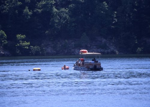 A boat on a calm lake with two people swimming nearby and a floating object in the water. Lush greenery surrounds the scene.