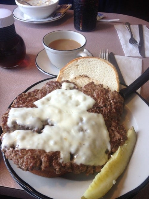A plate with a large piece of meat covered in white sauce, served with a slice of bread and a pickle on the side.