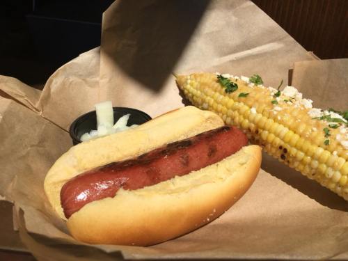 A hot dog in a bun with diced onions and a grilled corn on the cob topped with herbs and cheese, served on brown paper.