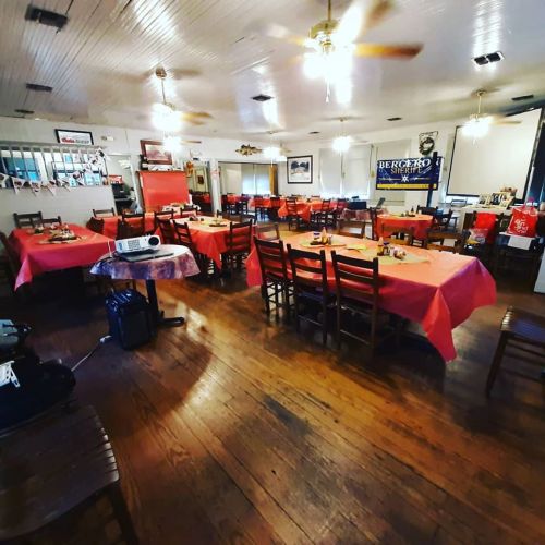 A spacious restaurant with wooden floors, tables covered in red cloths, and ceiling fans, set for dining.