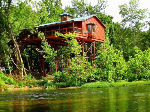 A red treehouse on stilts surrounded by lush green trees, overlooking a calm river.