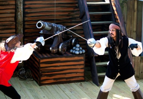 Two pirates duel with swords on a wooden deck, with a cannon and cannonballs in the background.