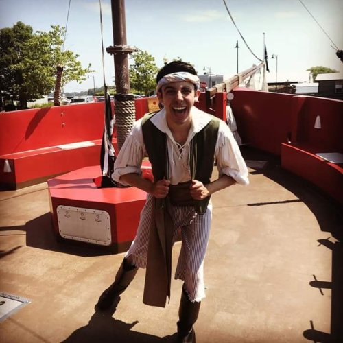 A smiling person in pirate attire stands on a red ship deck, striking a playful pose.