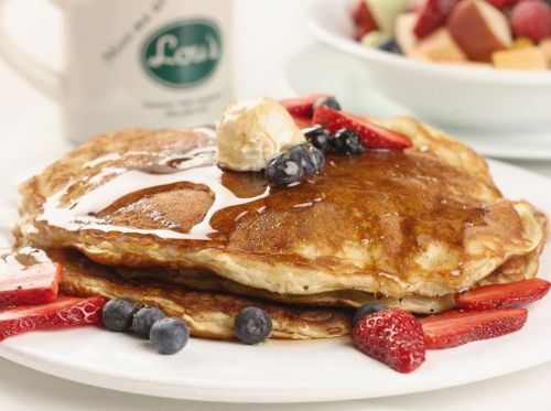Fluffy pancakes topped with butter, syrup, strawberries, and blueberries, served with a side of fresh fruit.