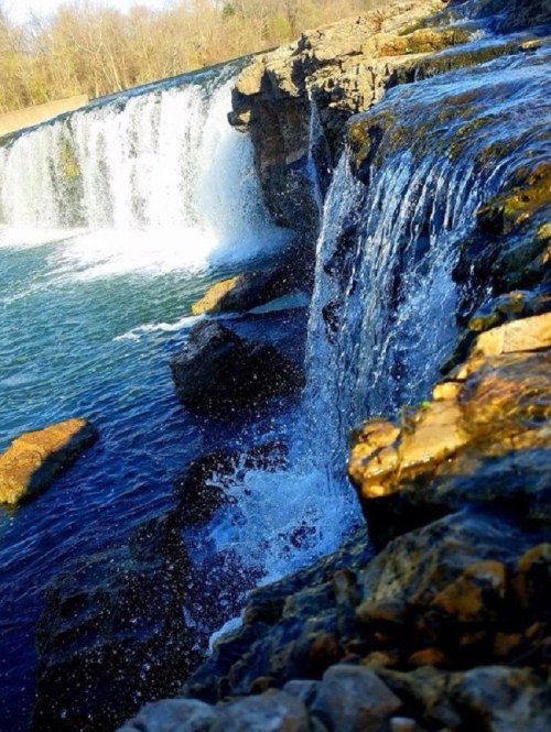 A scenic waterfall cascading over rocky cliffs into a clear blue pool, surrounded by trees and natural landscape.