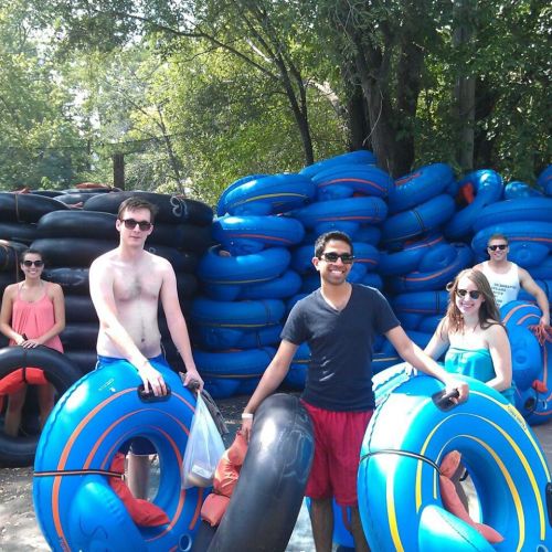 A group of five friends poses with colorful inner tubes near a stack of tubes in a sunny outdoor setting.