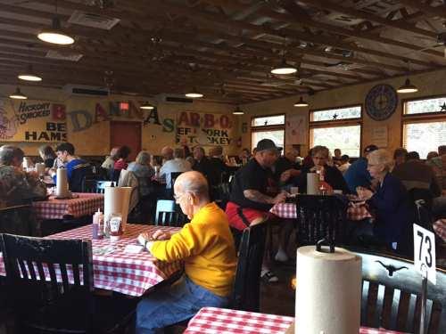 A busy restaurant interior with patrons dining at checkered tables, enjoying barbecue in a cozy, rustic setting.