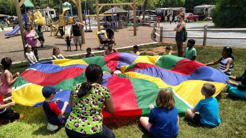 Children and adults play with a colorful parachute at a park, surrounded by playground equipment and a grassy area.