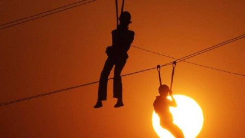 Two silhouetted figures ziplining against a vibrant sunset, with a large sun glowing in the background.