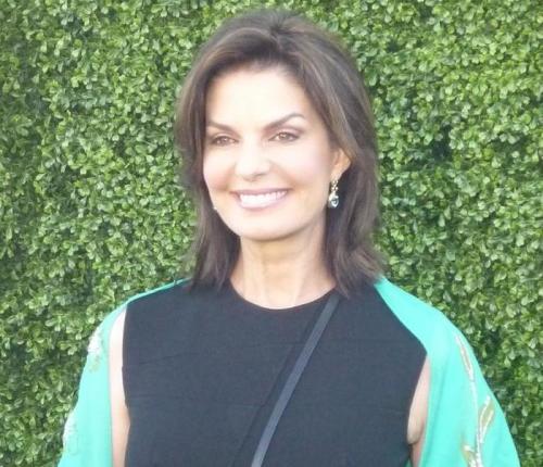 A woman with shoulder-length brown hair smiles, wearing a black dress with a turquoise shawl against a green backdrop.