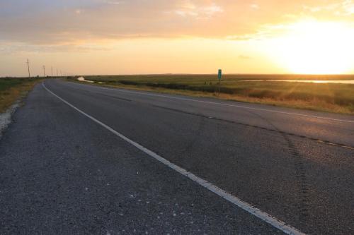 A winding road stretches into the distance under a colorful sunset, with fields and water visible on either side.