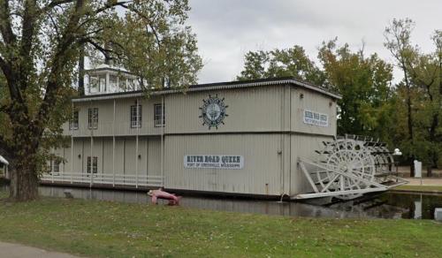 A large, white riverboat with a paddlewheel, docked near trees and a grassy area, labeled "River Road Queen."